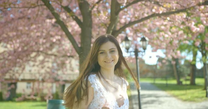 Happy smiling brunette is spinning round in the white pompous dress in the beautiful blooming sakura garden. 4k footage.