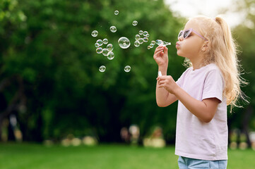 Cheerful caucasian blond preschool girl wearing sunglasses blowing soap bubbles in summer park...
