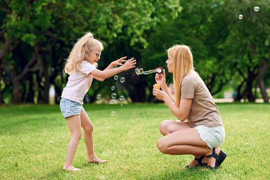 Preschool European laughing blond child girl with mother catching and bursting soap bubbles outdoors in summer park. Happy childhood concept