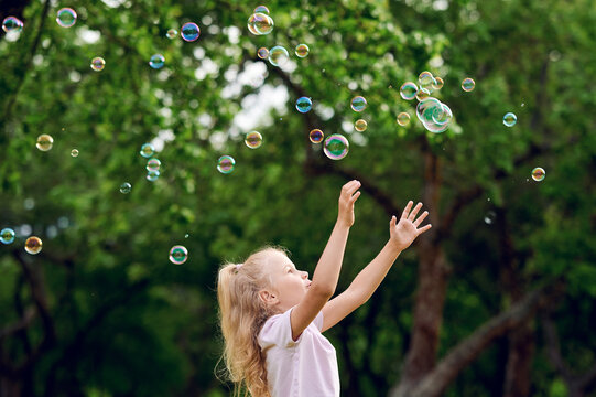 Preschool caucasian blond child girl catching soap bubbles outdoors in summer apple park. Happy childhood concept