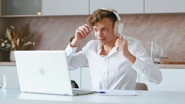 Man put headphones on and starts voice call on laptop. Young adult student participating in virtual conference call from kitchen. Employee on remote meeting from home laptop. Handheld shot.
