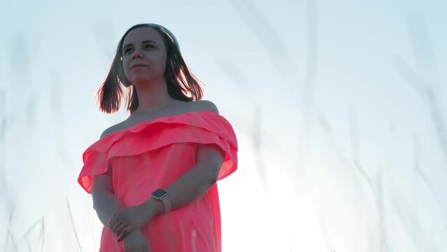 Young woman in bright dress with short hair, wireless headphones listening to music in windy weather against sky on eve of sunset.