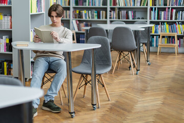 teenage guy sitting with book in reading room of library.