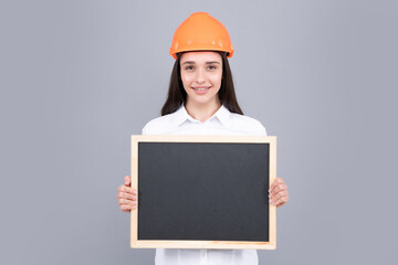 Young beautiful smiling architect woman in hard hat helmet hold board over gray background.