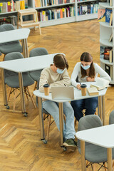 Fototapeta na wymiar high angle view of teenagers in medical masks writing near laptop and books in library.