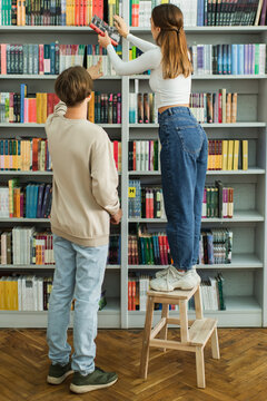 back view of teenage girl standing on step stool near friend and choosing books on shelves.