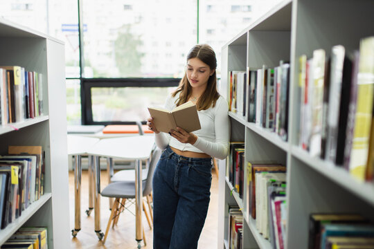 teenage girl standing near shelves in library and reading book.