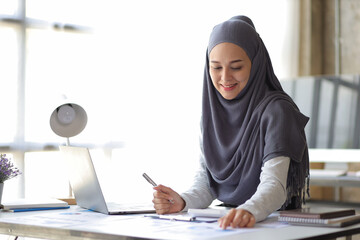 Attractive Muslim businesswoman working with laptop and analyzing financial statements on desk in...