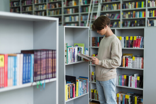 teenage student reading book near racks in library.