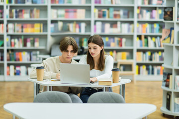 couple of teenagers studying near laptop and paper cups in library.