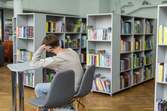 back view of teenage student sitting in library reading room near bookshelves.