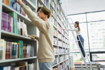 teenage girl standing on ladder and choosing books near friend on blurred foreground.