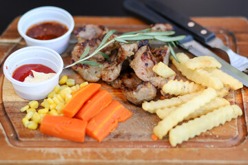 Grilled pork chop steak served with french fries, corn, carrot, ketchup, and Asian-style spicy sauce, on a wooden board
