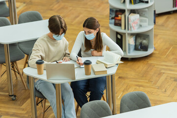 high angle view of teenage students in medical masks writing near laptop and paper cups in library.