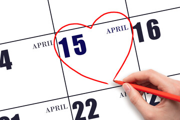 A woman's hand drawing a red heart shape on the calendar date of 15 April. Heart as a symbol of...