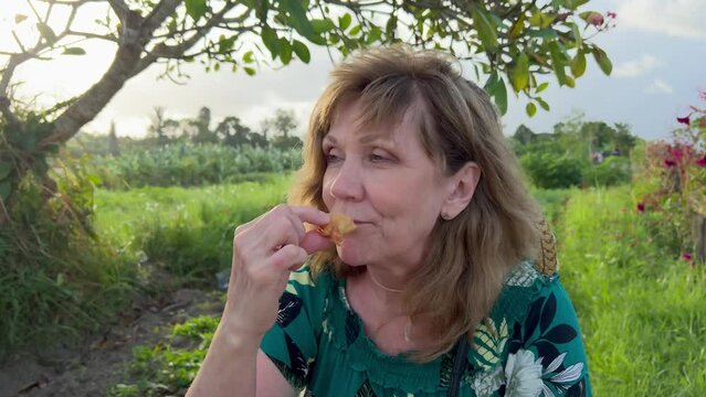 A beautiful mature caucasian woman eating a pastry in rural area enjoying summer picnic on a windy day at golden hour. Blonde female outdoors. Film grain. Soft focus. Live camera. Blur