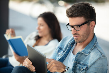 guy uses tablet next to the sitting girl reading book