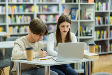 Fototapeta na wymiar teenage student writing in notebook near laptop and friend in reading room.
