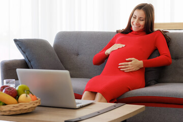 A beautiful, big pregnant woman sits on the sofa and uses her laptop computer to chat on a video call happily and comfortably. Family relationship and emotion care concept