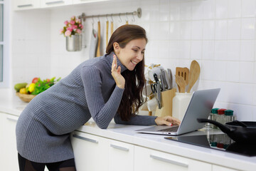Beautiful, cool, pregnant woman standing in modern kitchen and using laptop computer to chat in videocall with happy and relaxing manner - 527639575