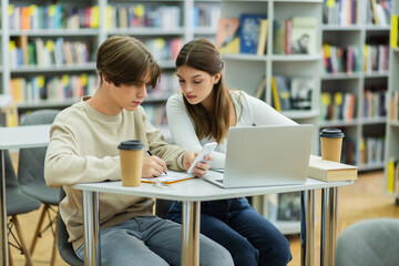 student holding smartphone and writing near laptop and teenage girl in library reading room.