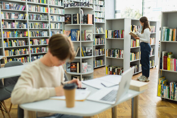 teen girl choosing books while her friend sitting near laptop on blurred foreground.