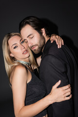 Seductive blonde woman hugging boyfriend in jacket on black background.