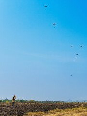 Back view of woman takes video clips and photo with tablet and watch with worry and concern during parachute training from airplane for army cadet in field with parachute and landscape in background.