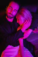 Young man hugging blonde girlfriend on leather couch with lighting isolated on black.