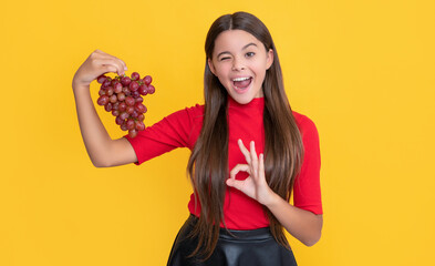 winking teen kid hold bunch of grapes on yellow background