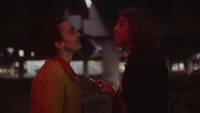 Waist up shot of two young female friends sharing earphones while listening to music, singing and dancing in red neon light under urban bridge