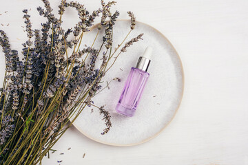 Obraz na płótnie Canvas Cosmetic oil or serum in purple bottle with dropper on a plate with bunch of lavender flowers on white table. Aromatherapy concept. Top view, flat lay