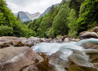 Paisaje alpino con un arroyo que discurre entre rocas y bosques con altas montañas de fondo de imagen.