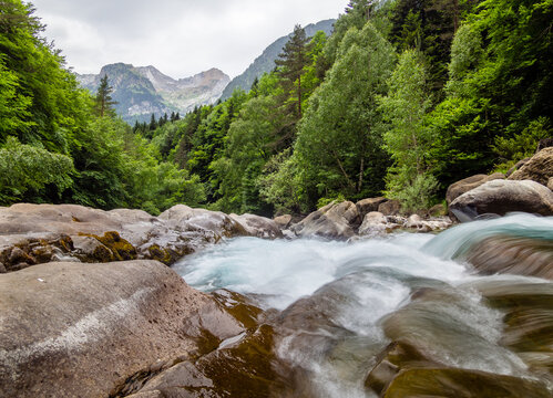 Paisaje alpino con un arroyo que discurre entre rocas y bosques con altas monta&ntilde;as de fondo de imagen.