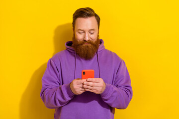 Photo of cute cheerful man wear violet sweatshirt typing modern device isolated yellow color...