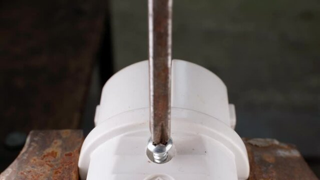 A Phillips screwdriver unscrews a screw from a white power plug close-up. The process of parsing the body of a plastic fork clamped in a vice. Repair and maintenance of electrical equipment
