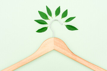 Wooden clothes hanger with green leaves over mint background as a symbol of conscious consumerism,...