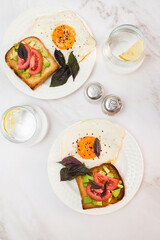 two plates of sandwiches with avocado, tomatoes, fried eggs, two glasses of water with lemon on light background.