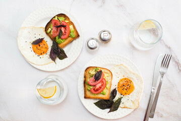 two plates of sandwiches with avocado, tomatoes, fried eggs, two glasses of water with lemon on light background.
