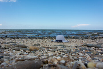 Fototapeta na wymiar A blue female hat and beach sunglasses lie on a sandy beach near the stones and an eye of sea waves, the concept of sea recreation and swimming in the sea.