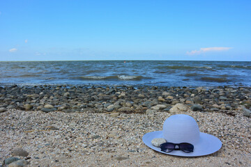 Fototapeta na wymiar A blue female hat and beach sunglasses lie on a sandy beach near the stones and an eye of sea waves, the concept of sea recreation and swimming in the sea.