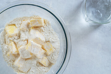 cubes cold butter  in a glass bowl with all-purpose flour in preparation to make puff pastry with a jar of ice water, basic puff pastry ingredients.