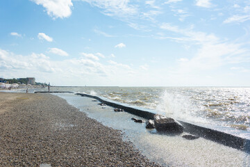 Waves splash the pier and rocks near the shore on a sunny and windy summer day on the sea coast.