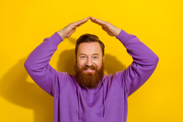 Photo of positive cute man wear violet sweatshirt showing arms roof space isolated yellow color background