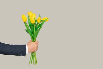A man's hand stretches forward a bouquet of flowers of yellow tulips on a gray background