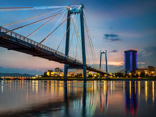 Suspension bridge bridge across the river at night. Night illumination of buildings, reflections.