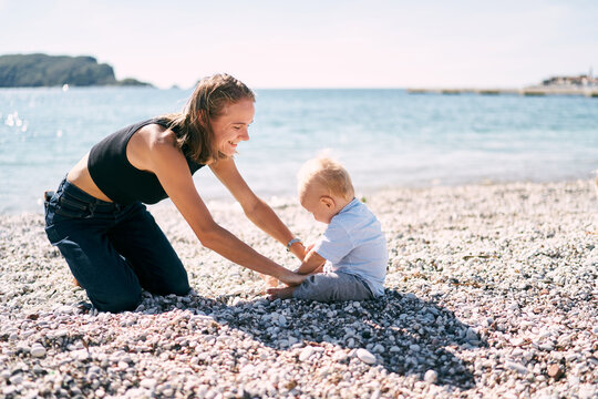 Mom sits in front of a little boy on a pebble beach and holds his hands. High quality photo