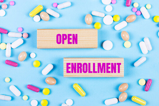 On a light blue background, multi-colored pills and wooden blocks with the text OPEN ENROLLMENT. Pharmaceutics. Medical concept.