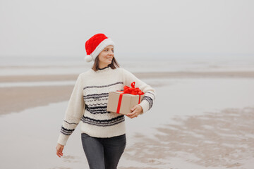 Obraz premium woman in a Santa Claus hat with a gift box in her hands smiles. a happy European woman carries a gift for New Year or Christmas. give gifts and surprises, poster postcard. holiday holidays Christmas