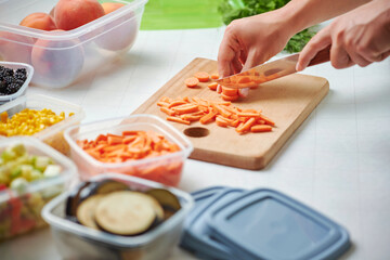 Hands of young female chopping fresh carrot on wooden board while preparing vegetables for freezing. Plastic containers with raw cut vegetables for freezing on kitchen table